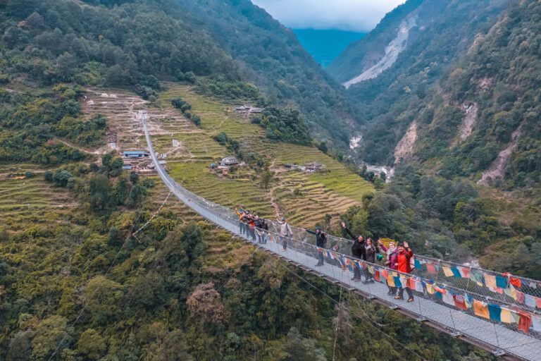 Eine Gruppe von WeRoad-Reisenden winkt von einer langen Hängebrücke über einem tiefen Tal im Himalaya in Nepal.