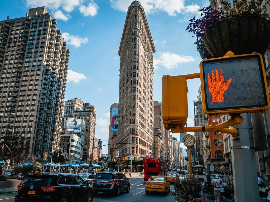 Blick auf das Flatiron Building in New York City bei Tageslicht mit Verkehr und Fußgängern im Vordergrund.