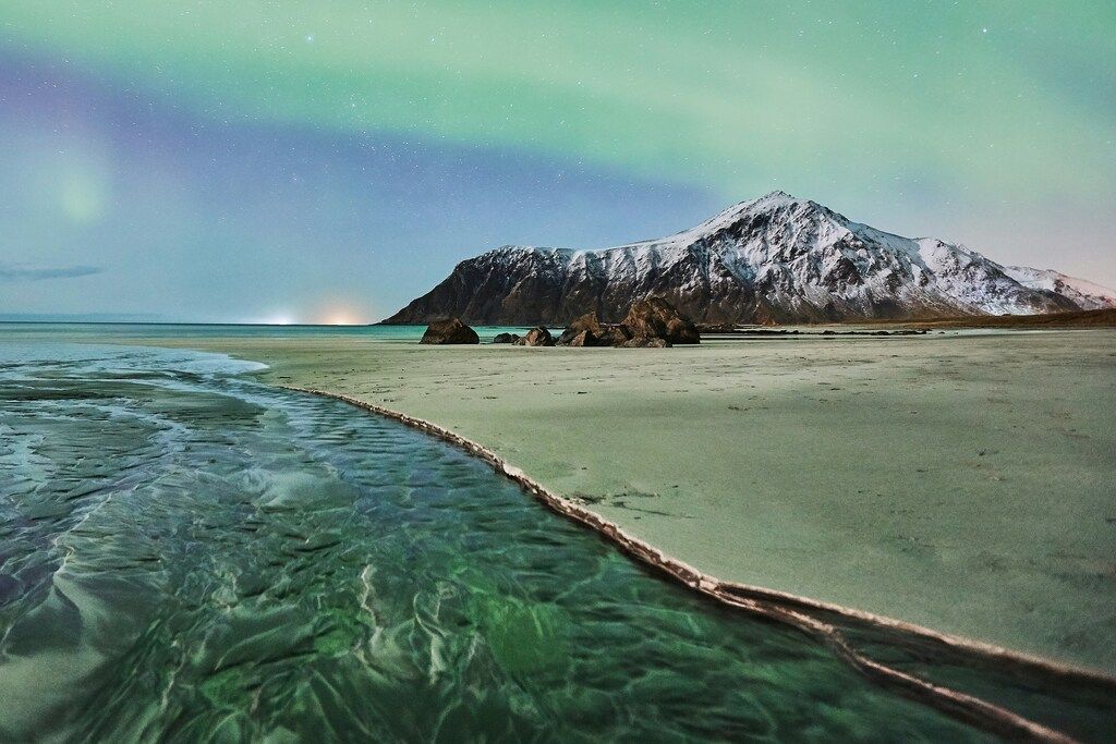 Grüne Nordlichter über einem Strand mit Bergen im Hintergrund.