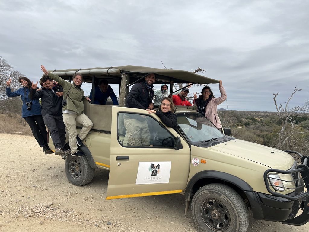 Eine Gruppe von WeRoad-Reisenden lächelt und winkt aus einem Safari-Geländewagen in der südafrikanischen Savanne unter einem bewölkten Himmel.
