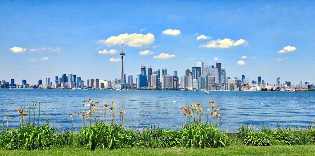 Panoramablick auf die Skyline von Toronto mit dem CN Tower im Hintergrund, gesehen von einem grünen Ufer.
