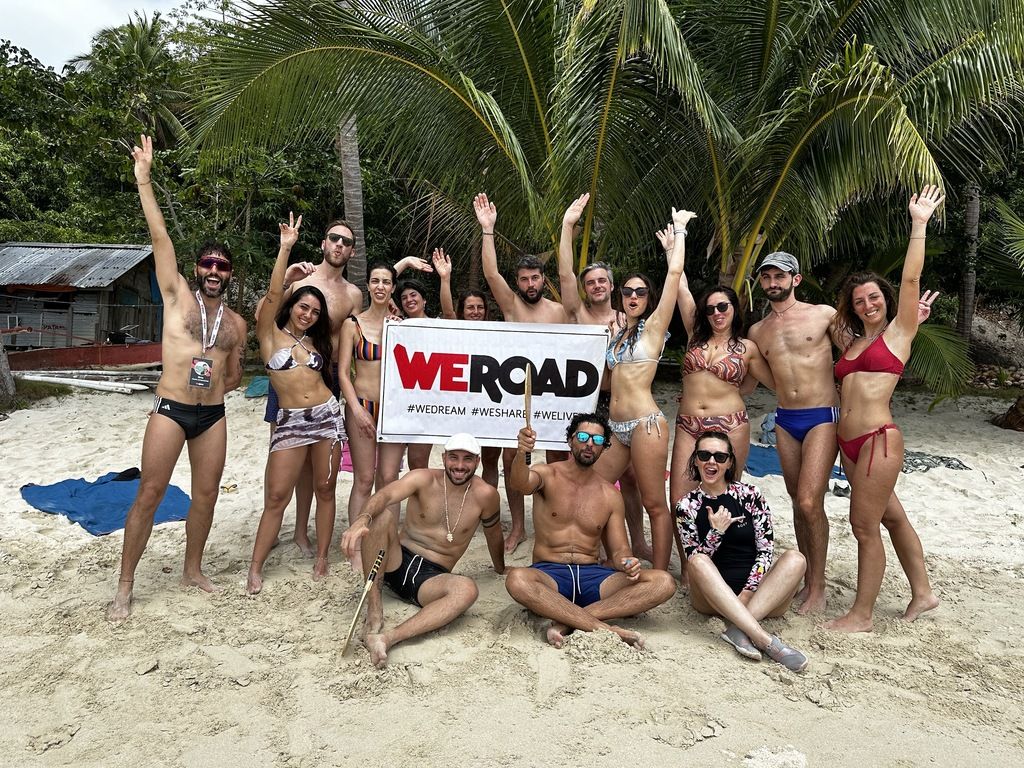 Eine Gruppe von WeRoad-Reisenden in Badekleidung posiert fröhlich mit einem Banner an einem tropischen Strand auf den Philippinen.