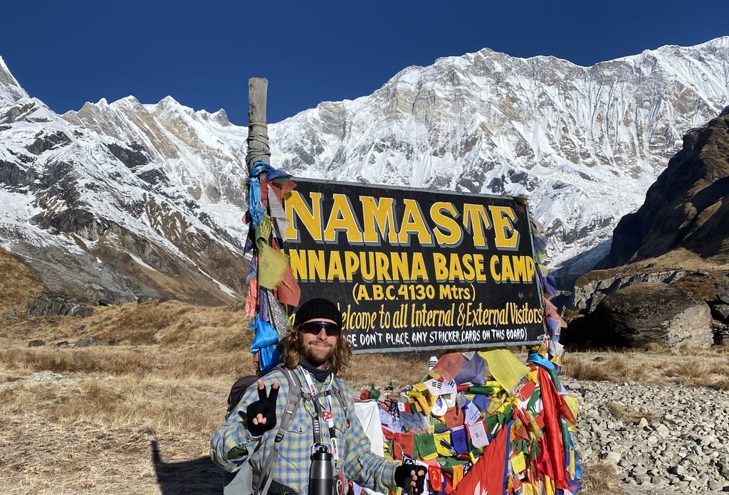 Ein WeRoad-Reisender posiert mit dem Friedenszeichen vor dem 'Namaste Annapurna Base Camp' Schild im nepalesischen Himalaya.