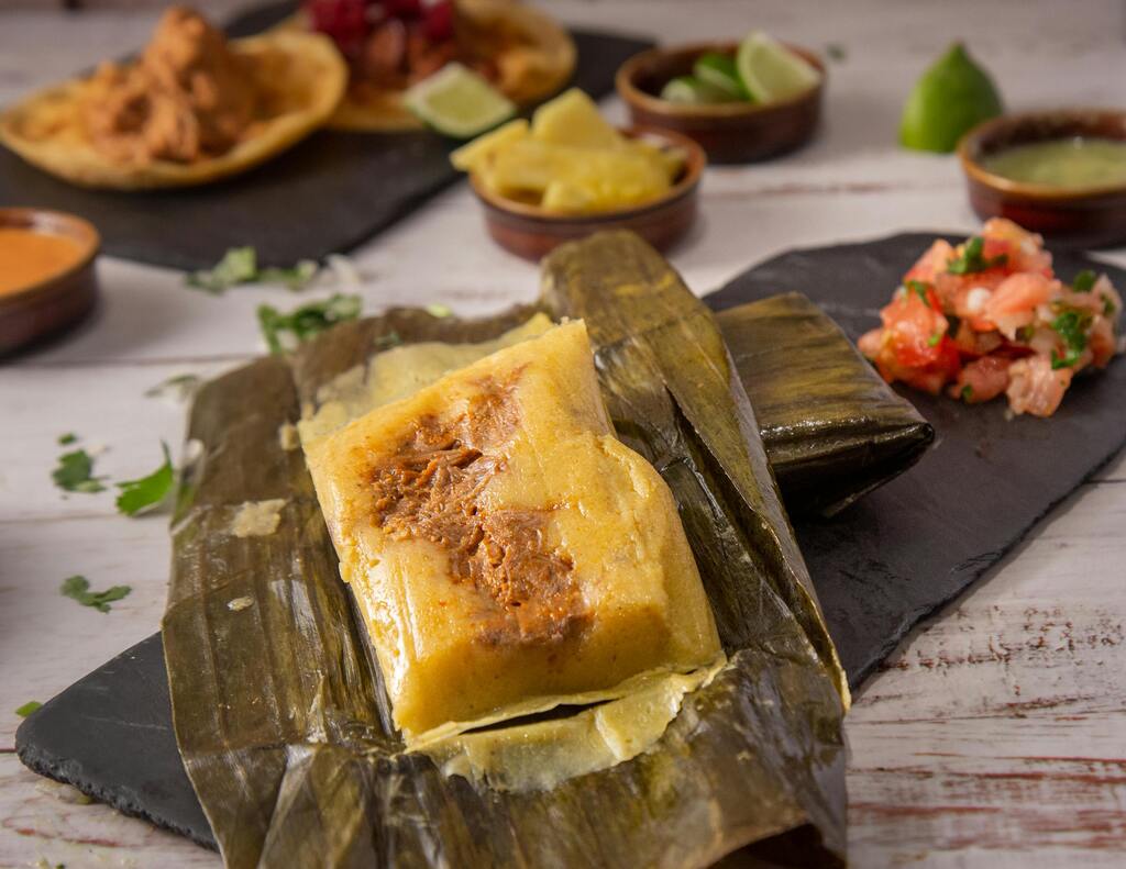 Tamales mit Fleischfüllung in Bananenblättern serviert, begleitet von Beilagen