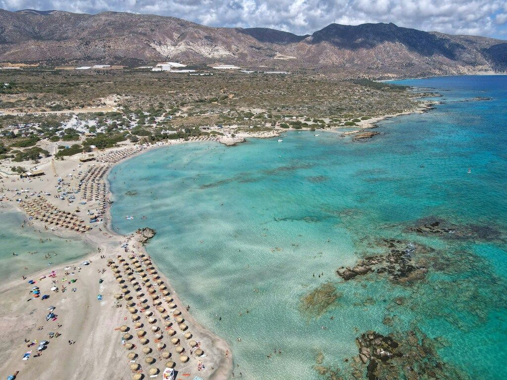 Luftaufnahme des Strandes von Elafonissi auf Kreta mit türkisfarbenem Wasser und Sonnenschirmen in Reihen.