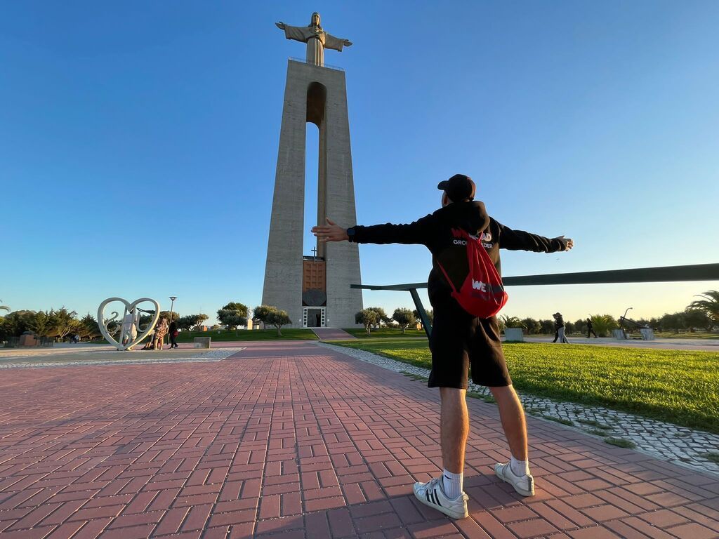 WeRoad-Reisender mit ausgestreckten Armen vor der Cristo-Rei-Statue in Lissabon bei Sonnenuntergang.