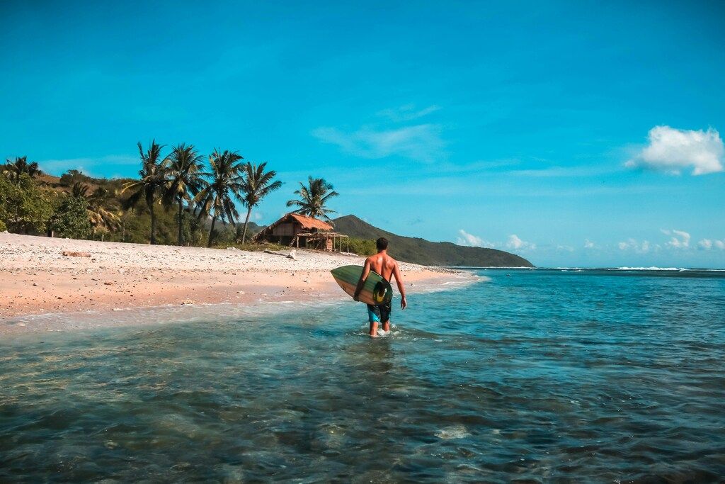Surfer geht mit Brett ins türkisfarbene Meer vor einer tropischen Strandlandschaft mit Palmen