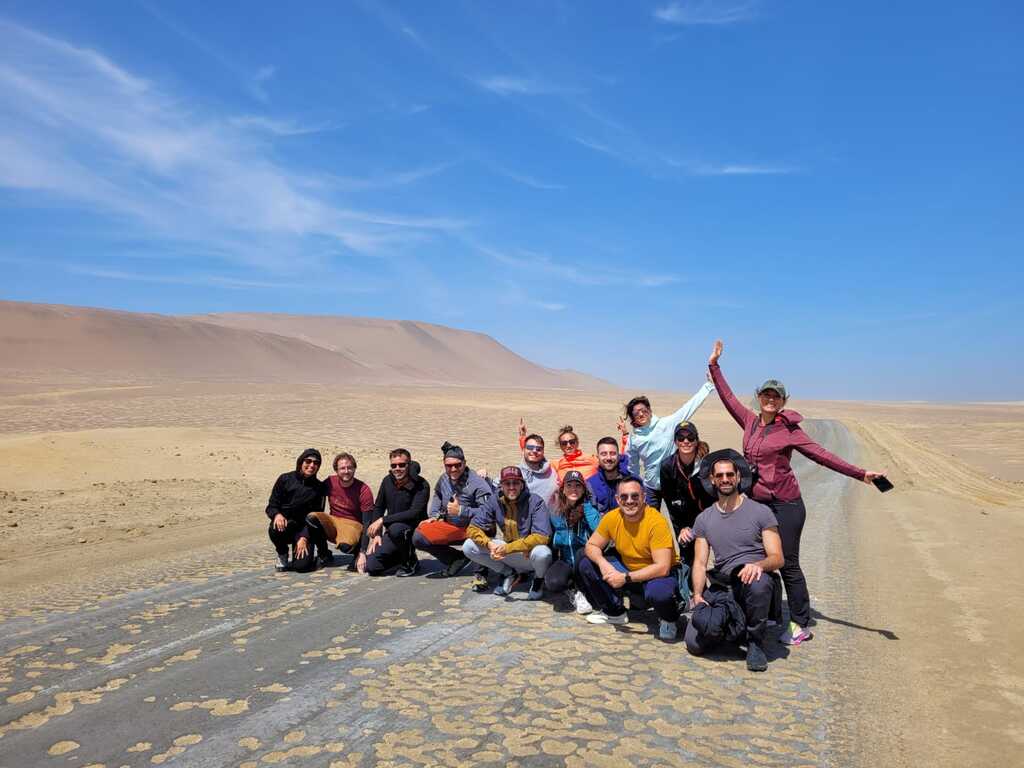 Gruppe von WeRoad-Reisenden posiert lachend auf einer Straße inmitten der peruanischen Wüste unter blauem Himmel.