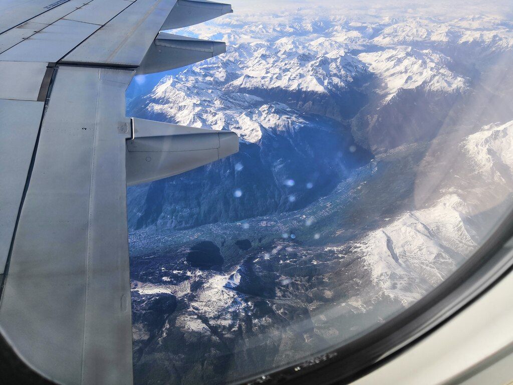 Luftaufnahme der verschneiten Alpen durch das Fenster eines Flugzeugs mit Flügel im Vordergrund.