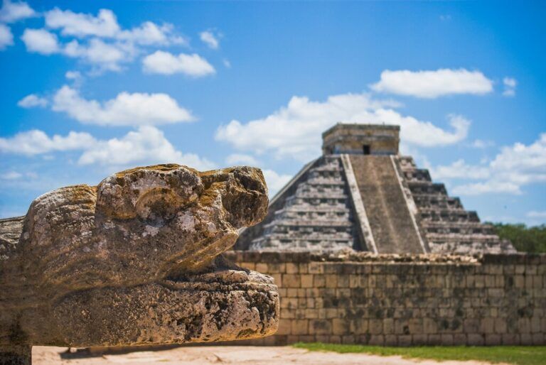 Steinskulptur eines Kopfes einer gefiederten Schlange (Kukulkan) mit der Pyramide von Chichén Itzá im Hintergrund, unter blauem Himmel in Mexiko.