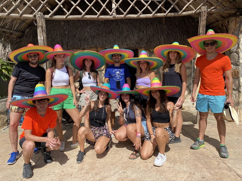 WeRoad-Reisegruppe trägt bunte Sombreros vor einer traditionellen Hütte in Mexiko.