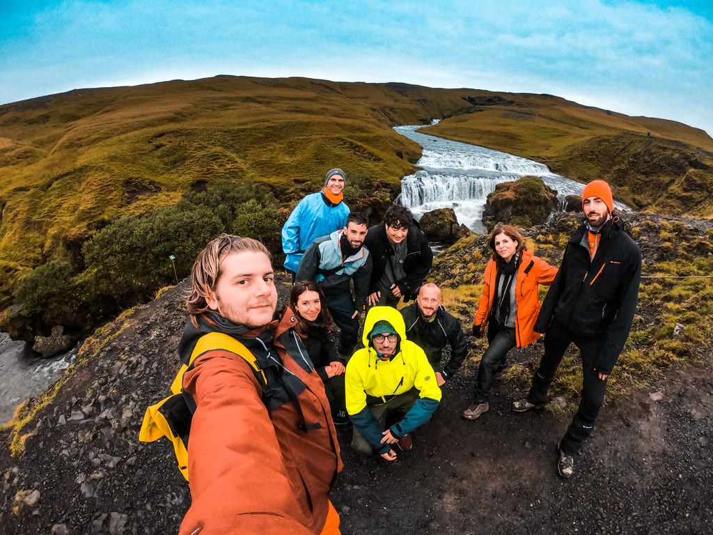 WeRoad-Reisende in wetterfester Kleidung machen ein Gruppenfoto vor einem Wasserfall in der grünen Landschaft Islands.