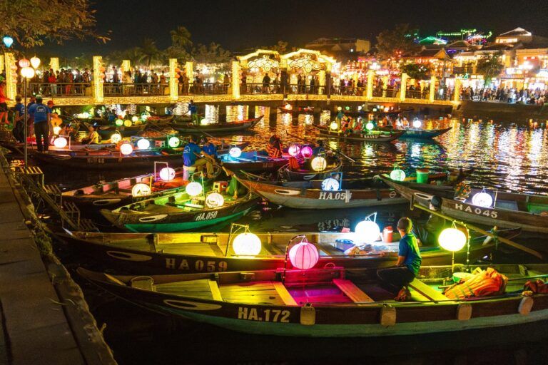 Boote mit leuchtenden Laternen auf dem Fluss in Hoi An bei Nacht, mit einer beleuchteten Brücke im Hintergrund.
