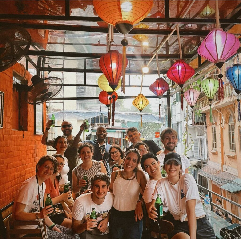Eine Gruppe von WeRoad-Reisenden lächelt und stößt mit Bierflaschen in einem vietnamesischen Lokal an, das mit bunten Lampions geschmückt ist.