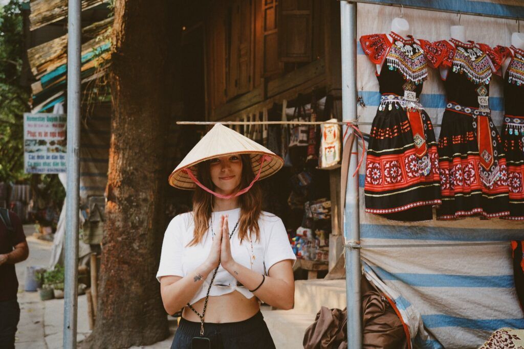 Reisende mit Kegelhut (Nón Lá) und aneinandergelegten Händen vor einem Stand mit Kleidern.
