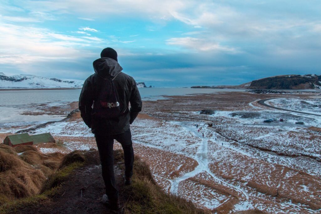 Reisender mit Rucksack blickt auf weite isländische Winterlandschaft mit Küste und Bergen.