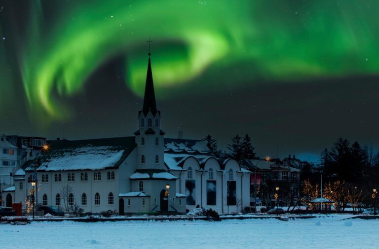 Die Nordlichter (Aurora Borealis) tanzen grün über einer kleinen, weißen Kirche in Reykjavik im winterlichen Schnee.