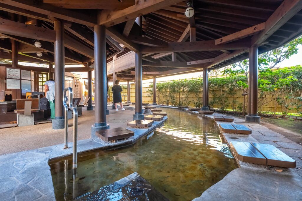 Überdachtes japanisches Ashiyu (Fußbad) mit hölzernen Sitzplatten und Besuchern.