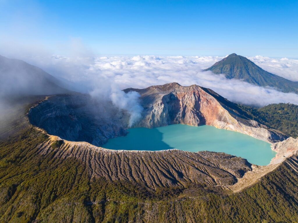 Türkisblauer Säuresee und rauchender Kraterrand des Ijen Vulkans auf Java über einem Wolkenmeer.