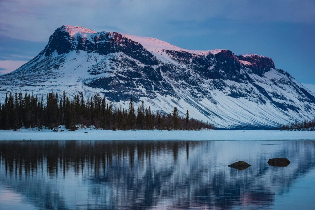 Majestätischer, schneebedeckter Fjäll-Berg, dessen Gipfel sich in einem ruhigen Bergsee spiegelt.