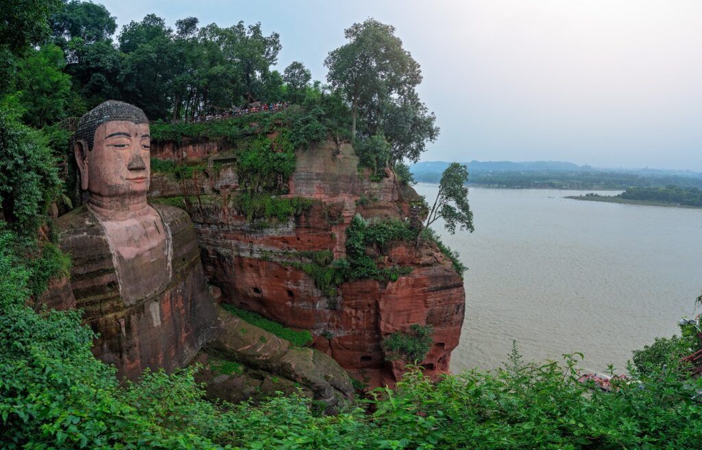 Der riesige Leshan Buddha an einer roten Felswand, umgeben von Vegetation, blickt auf den Fluss.
