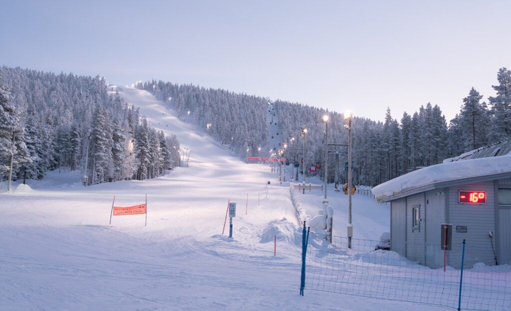 Beleuchtete Skipiste in Lappland, umgeben von verschneiten Nadelwäldern bei Dämmerung und sehr kalter Temperatur (-16°C).