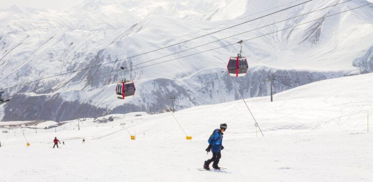 Snowboarder fährt eine breite, präparierte Piste hinunter. Im Hintergrund sind tief verschneite Berge und rote Gondeln der Seilbahn zu sehen.