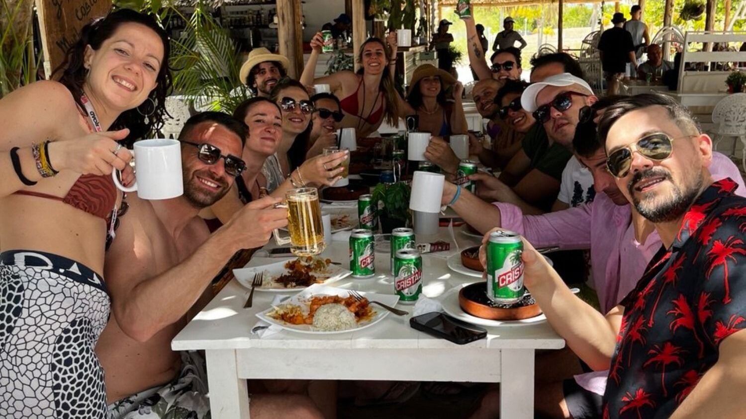 Eine gut gelaunte WeRoad-Reisegruppe stößt an einem weißen Holztisch in einem Strandrestaurant unter einem Strohdach mit Bier und Kaffee an.