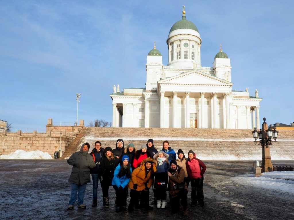 Eine WeRoad-Reisegruppe in Winterkleidung posiert lächelnd auf dem schneebedeckten Senatsplatz in Helsinki vor der imposanten weißen Domkirche.