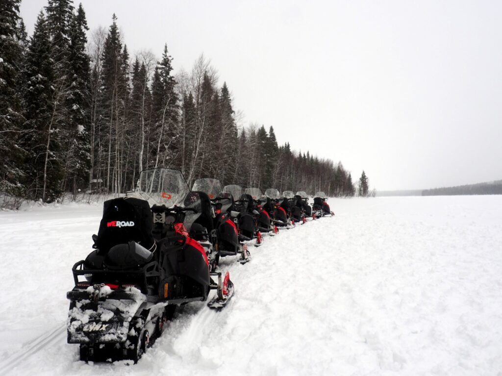Eine lange Reihe von Motorschlitten steht startbereit auf einem verschneiten Pfad in Rovaniemi, gesäumt von einem dichten Nadelwald unter einem grauen Winterhimmel.