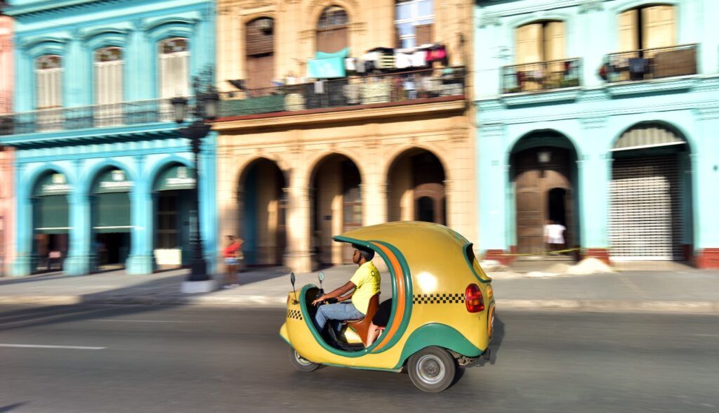 Ein markantes gelbes Coco-Taxi fährt an farbenfrohen Kolonialgebäuden in einer sonnigen Straße in Havanna.