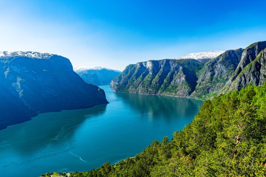 Blick von oben auf den weiten, blauen Sognefjord in Norwegen, umgeben von steilen, grünen Bergen mit schneebedeckten Gipfeln unter klarem Himmel.