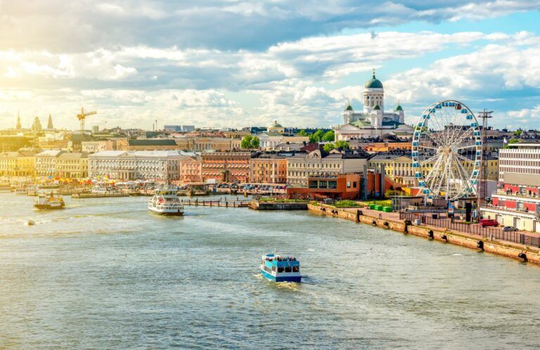 Eine sonnige Luftaufnahme des Hafens von Helsinki mit Blick auf die Domkirche, das Riesenrad 'SkyWheel' und mehrere Fähren, die auf dem glitzernden Wasser fahren.