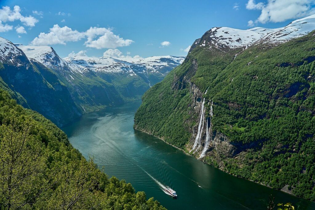 Ein Kreuzfahrtschiff fährt durch den tiefblauen Geirangerfjord in Norwegen, vorbei an der berühmten Wasserfallgruppe ‚Die Sieben Schwestern‘ und schneebedeckten Gipfeln.