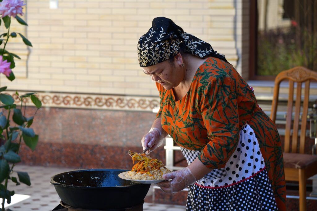Eine Frau in traditioneller Kleidung serviert eine Portion Plov aus einem großen Kessel im Freien.