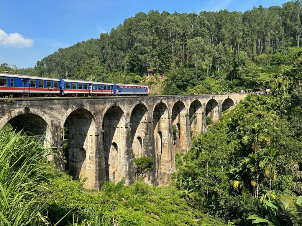 Ein blau-roter Zug überquert eine hohe Steinbogenbrücke inmitten einer üppigen, grünen Hügellandschaft unter blauem Himmel.