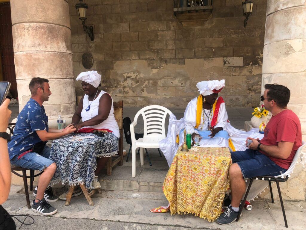 Zwei Reisende sitzen an Tischen im Freien bei zwei einheimischen Frauen in traditioneller weißer Kleidung in Havanna.
