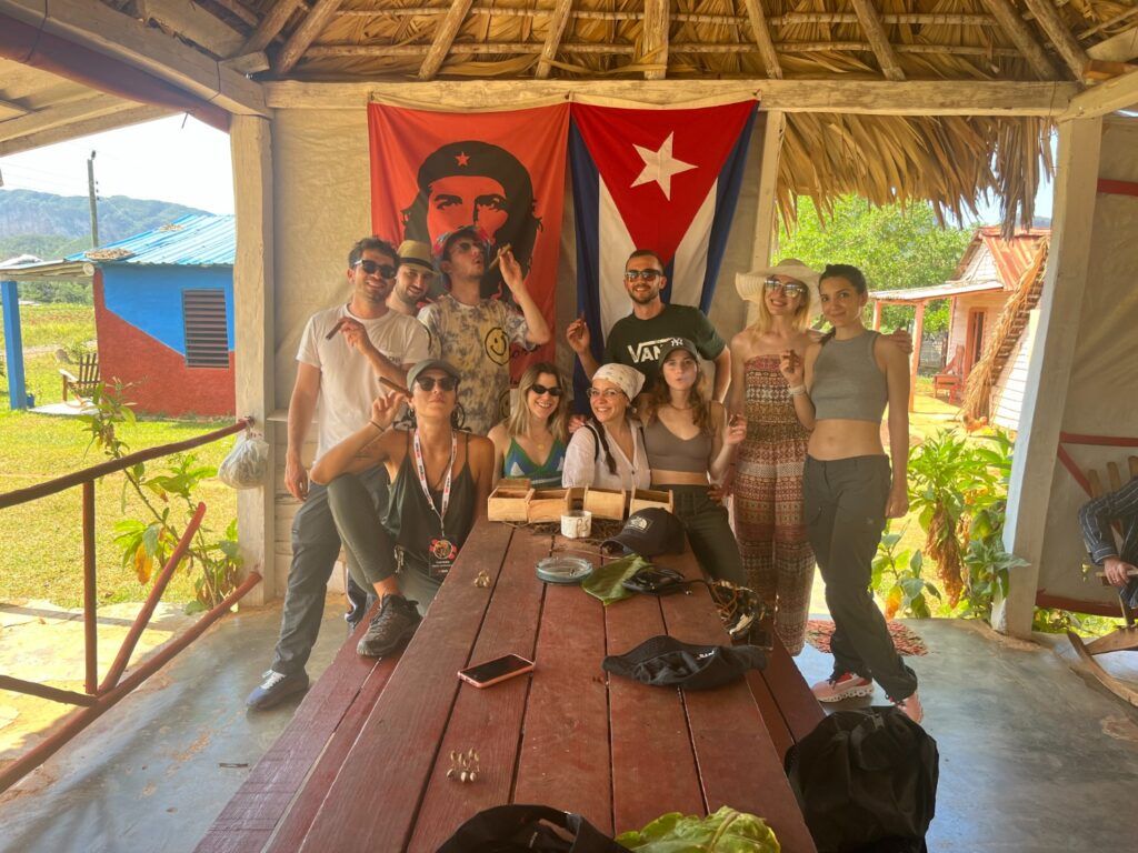 Eine Gruppe von WeRoad-Reisenden posiert lächelnd in einer rustikalen Holzhütte in Viñales, Kuba, vor einer kubanischen Flagge und einem Porträt von Che Guevara.