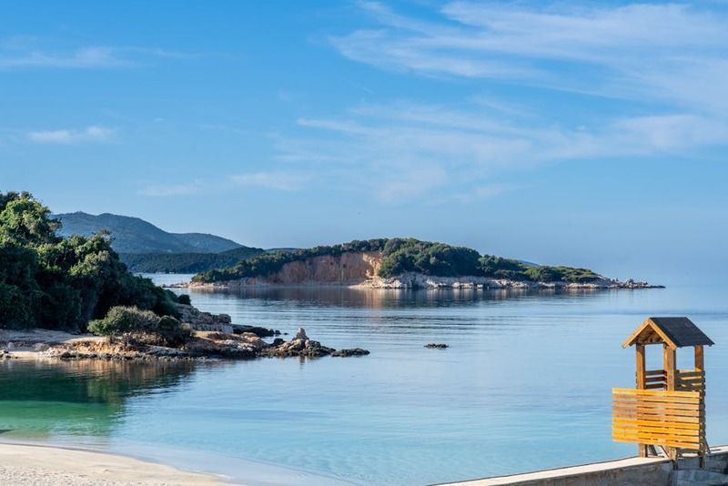Ruhige Küstenbucht mit kristallklarem türkisem Wasser, Felseninsel und gelbem Holzwächterturm in Albanien