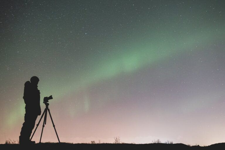 Silhouette eines Fotografen mit Stativ, der das gruene Nordlicht unter einem Sternenhimmel fotografiert