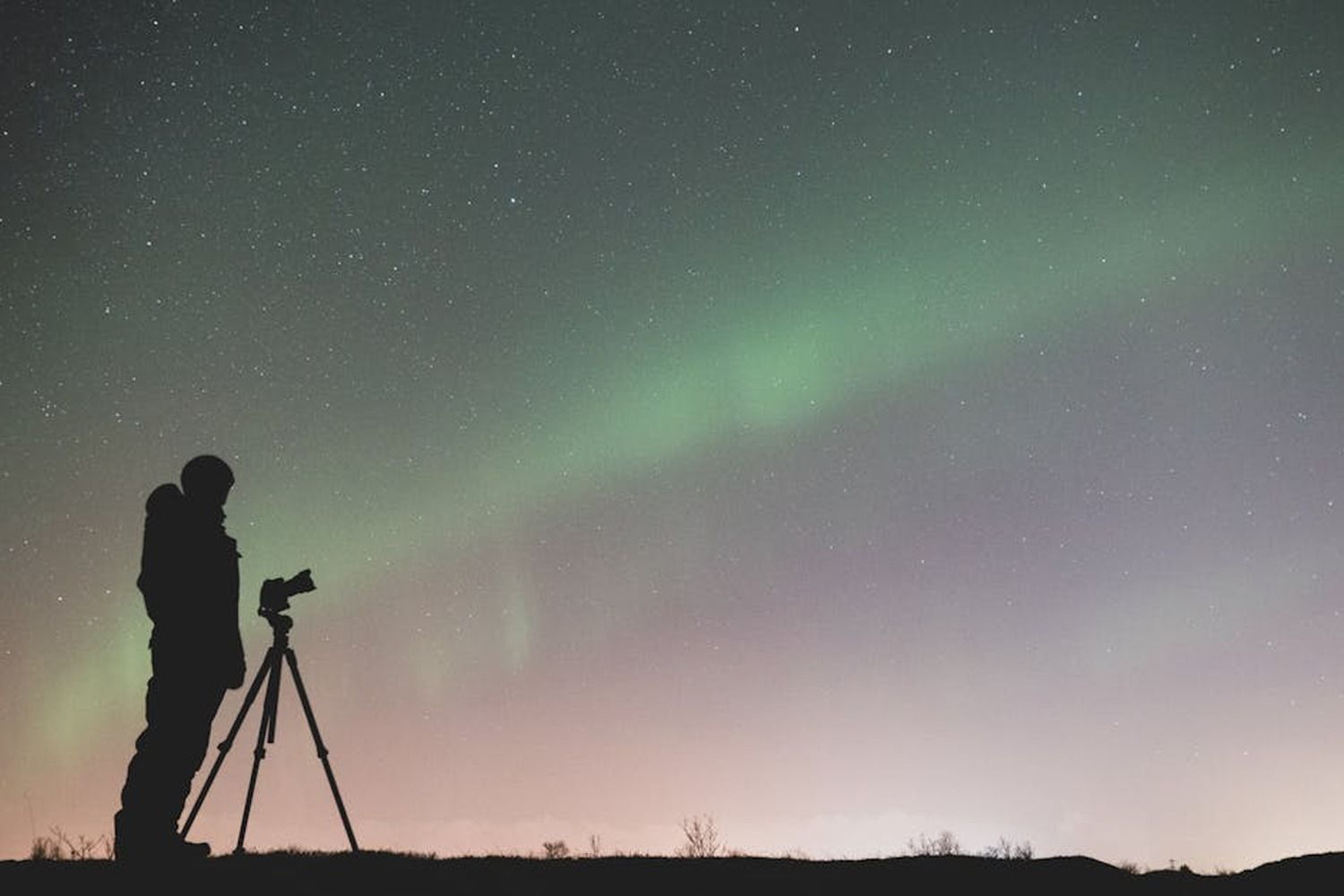 Silhouette eines Fotografen mit Stativ, der das gruene Nordlicht unter einem Sternenhimmel fotografiert