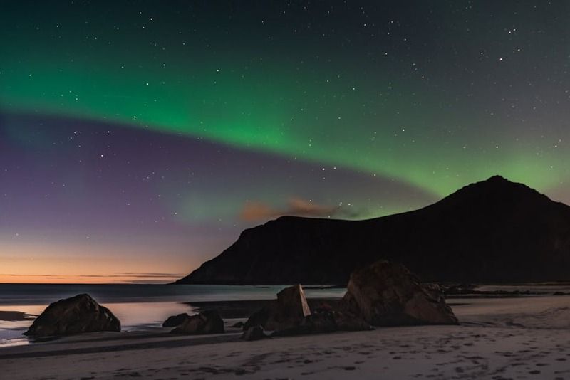 Nordlicht über einem Sandstrand mit dunklem Bergsilhouetten und farbigem Abendhimmel auf den Lofoten in Norwegen