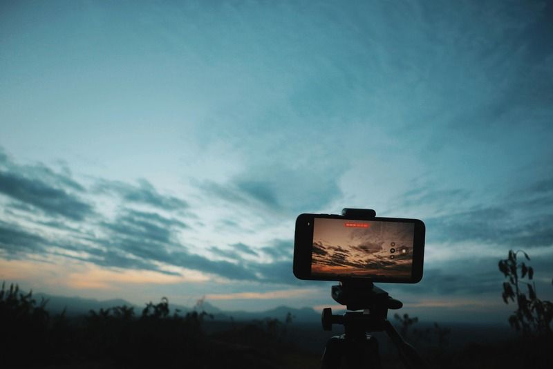 Smartphone auf Stativ nimmt einen bewoelkten Sonnenuntergang ueber einer Berglandschaft auf