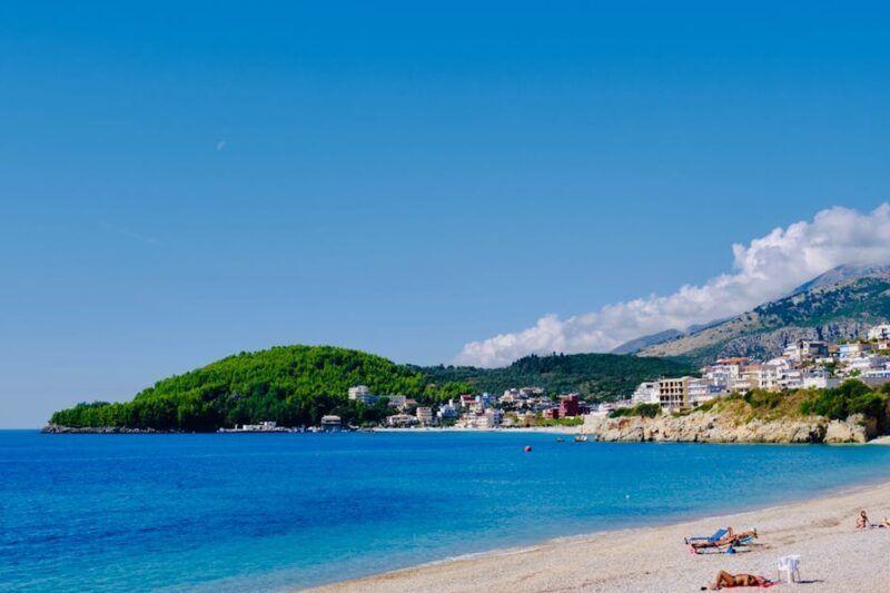 Panoramablick auf einen weißen Sandstrand mit türkisblauem Meer und Küstenstadt am Berghang in Albanien Panoramablick auf einen weißen Sandstrand mit türkisblauem Meer und Küstenstadt am Berghang in Albanien