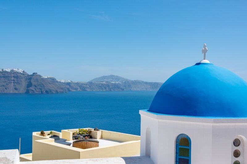 Ikonische blaue Kuppelkirche in Oia, Santorini, mit weißer Architektur und dem tiefblauen Wasser der Ägäis und den Vulkanklippen im Hintergrund Ikonische blaue Kuppelkirche in Oia, Santorini, mit weißer Architektur und dem tiefblauen Wasser der Ägäis und den Vulkanklippen im Hintergrund