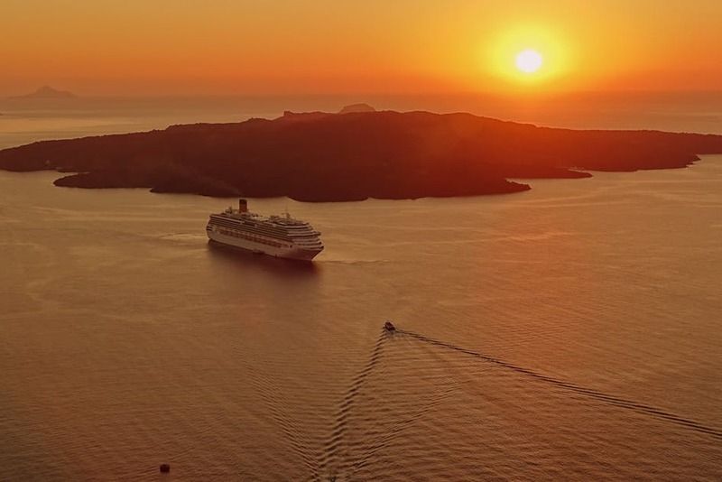 Kreuzfahrtschiff bei Sonnenuntergang auf dem Wasser der Santorini-Kaldera mit der Silhouette von Nea Kameni und orangerotem Abendhimmel