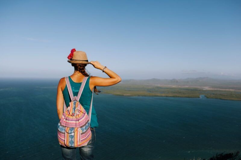 Rückenansicht einer Touristin mit Rucksack, die ihren Strohhut streichelt, während sie vom Berg aus das Meer betrachtet.