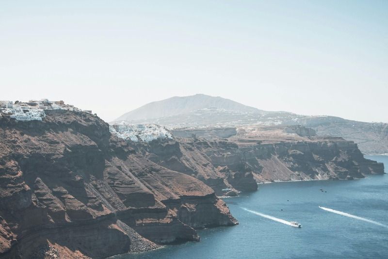 Blick von den Felsen der Santorini-Kaldera auf das tiefblaue Meer mit einem Motorboot und den weißen Häusern von Imerovigli in der Ferne