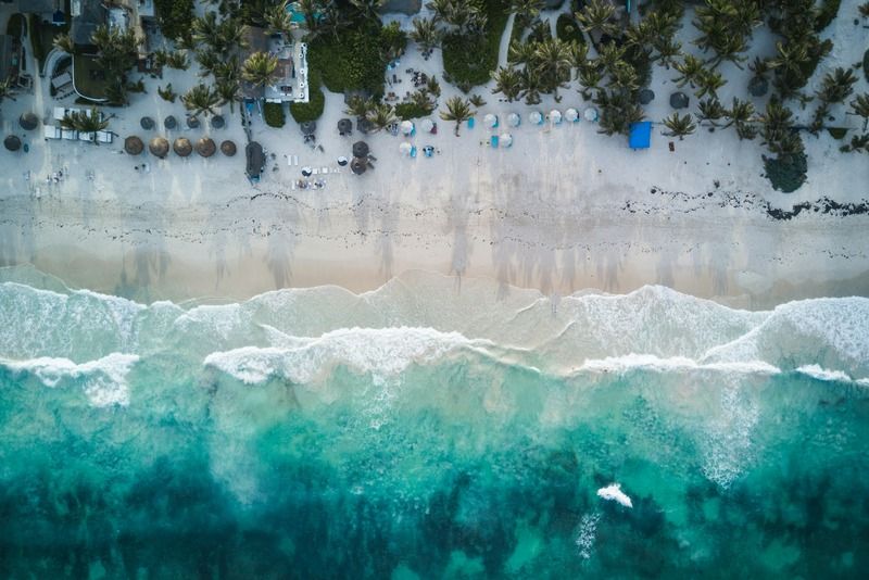 Drohnenaufnahme aus der Vogelperspektive auf den Strand von Tulum mit türkisfarbenen Wellen, Sonnenschirmen und Palmen, Mexiko