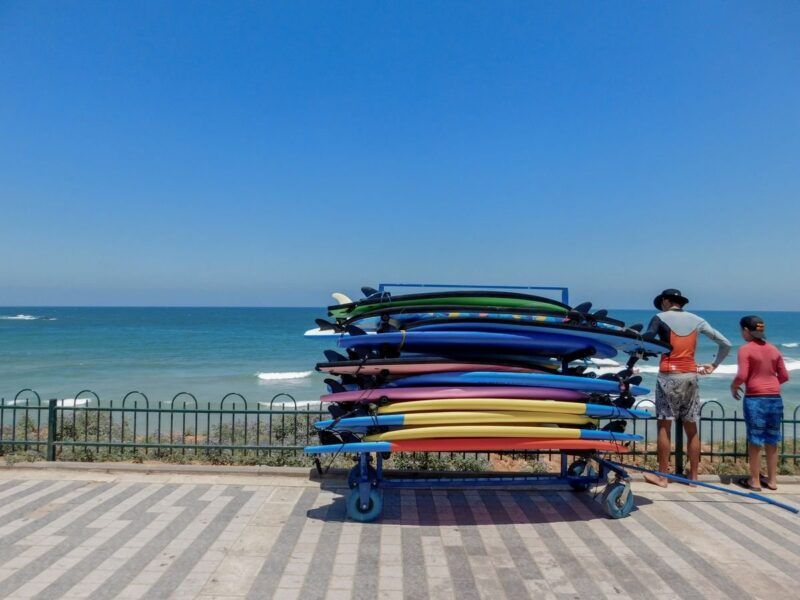 Bunter Stapel Surfboards auf einem Wagen an der Strandpromenade, daneben zwei Personen am Meer.